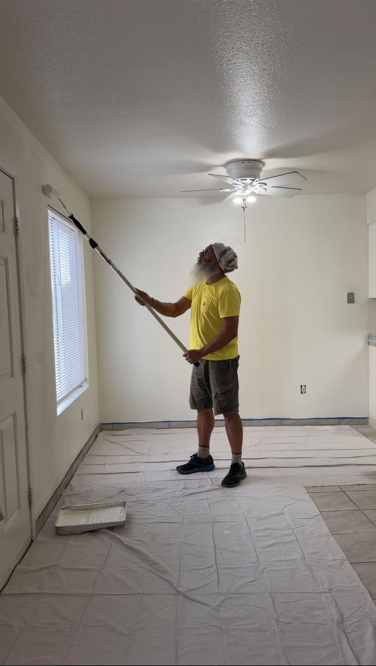 Painting a wall with a long-handled roller while protective sheets cover the apartment floor