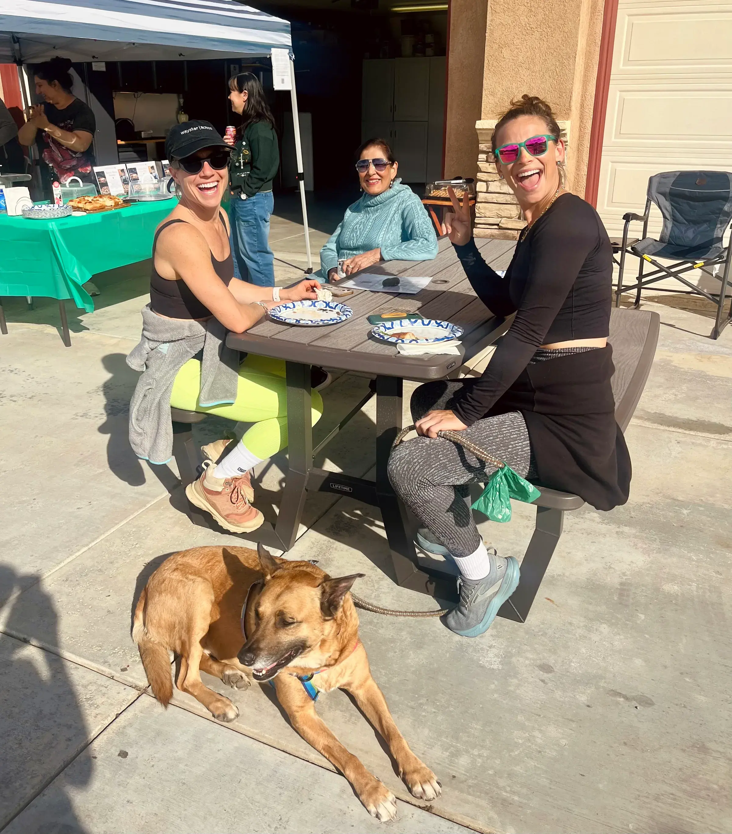 Three Pizza for Paws guests smiling around an outdoor table with a dog resting nearby